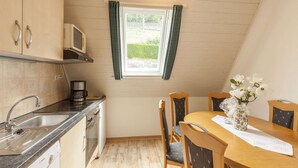 Una cucina compatta con forno a microonde, lavello, lavastoviglie Bosch e mobili in legno. Si nota un tavolo con sedie e un vaso di fiori. Si vede una finestra con tende, e la stanza ha un soffitto a falde.