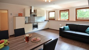 A modern kitchen with white cabinets, a dining table with a fruit bowl and a glass of beer, and a black sofa with teal cushions. - Modern Apartment in Eslarn With Lake Near (Eslarn)