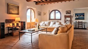 A cozy living room with a sofa, coffee table, and a fireplace. There are two windows with arched tops, a painting on the wall, and a bookshelf. - Beautiful Villa in San Giovanni del Pantano With Pool (Perugia)