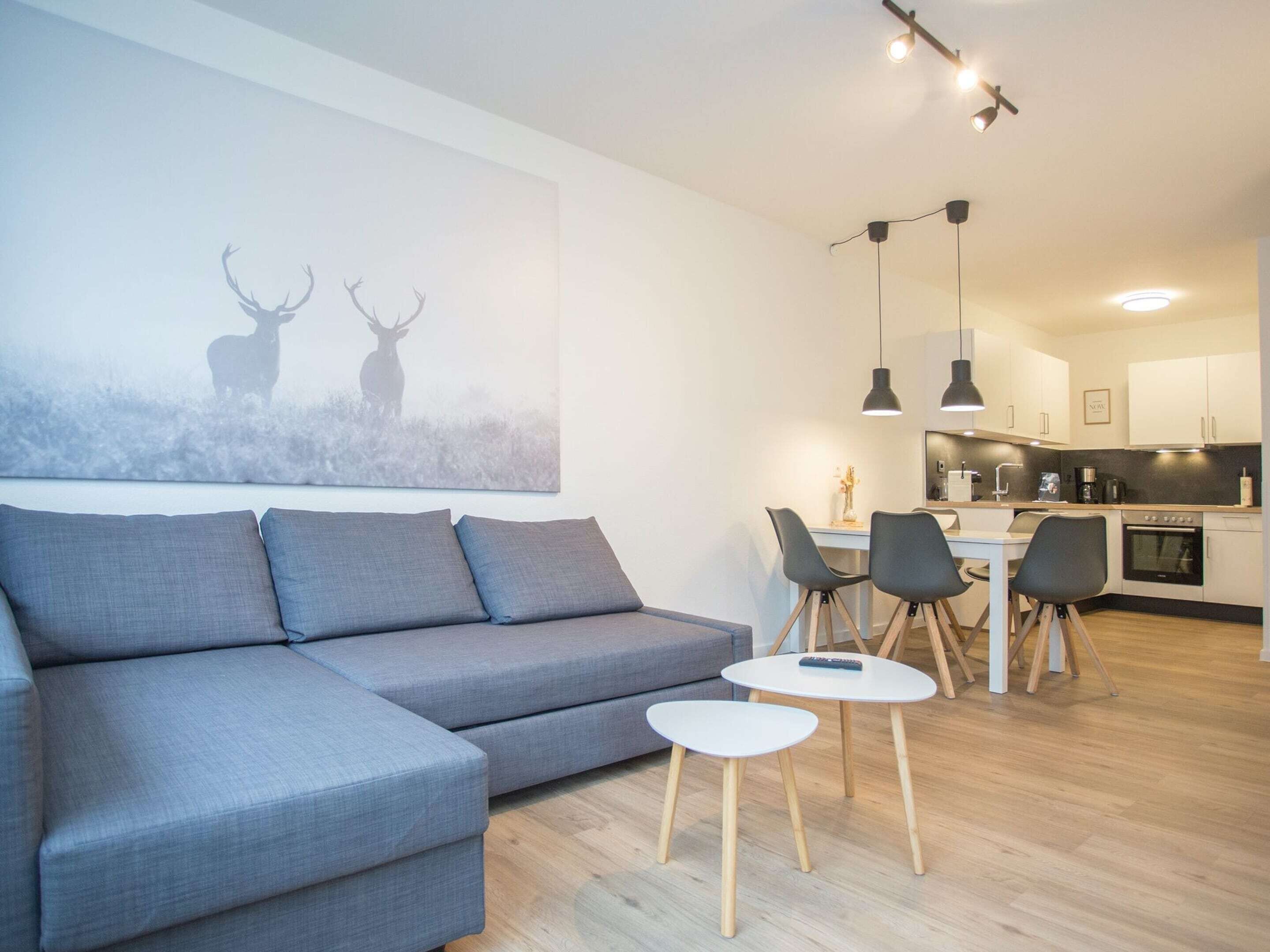 A modern living room with a grey sectional sofa, a small white table, and a dining area with chairs and a kitchen in the background.