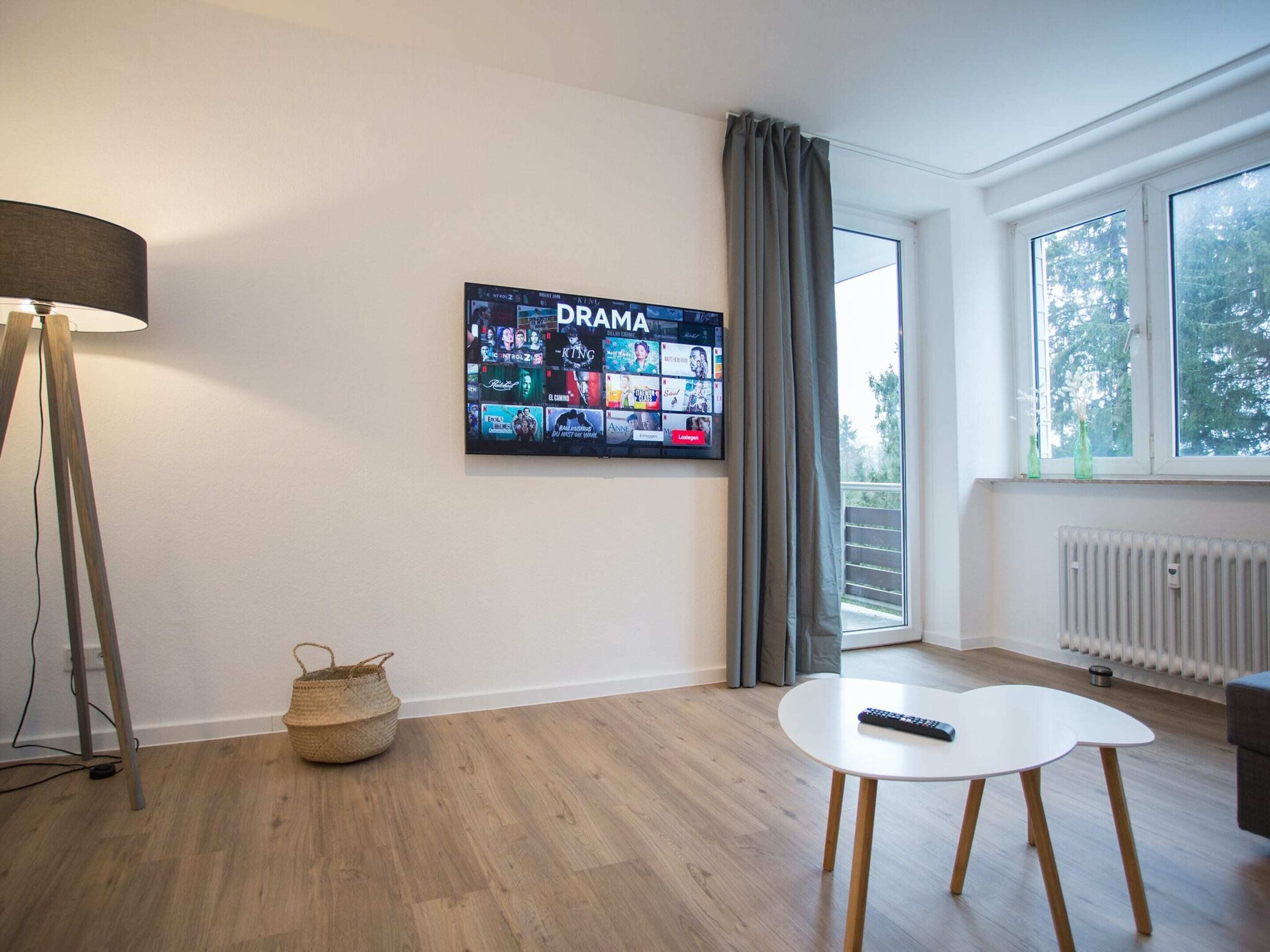 Un soggiorno moderno con televisione a schermo piatto che mostra un canale televisivo drammatico, una lampada da terra, un tavolino con un telecomando e vista sull'esterno attraverso una finestra.