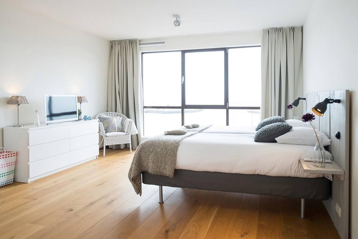 Un dormitorio moderno con un ventanal grande, una cama con almohadas, una mesita de noche con lámparas, una silla y un aparador.
