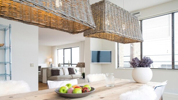 Een moderne woonkamer met een houten eettafel, zachte witte stoelen, een schaal fruit, rieten plafondlampen, een flatscreen televisie en een groot raam met jaloezieën.