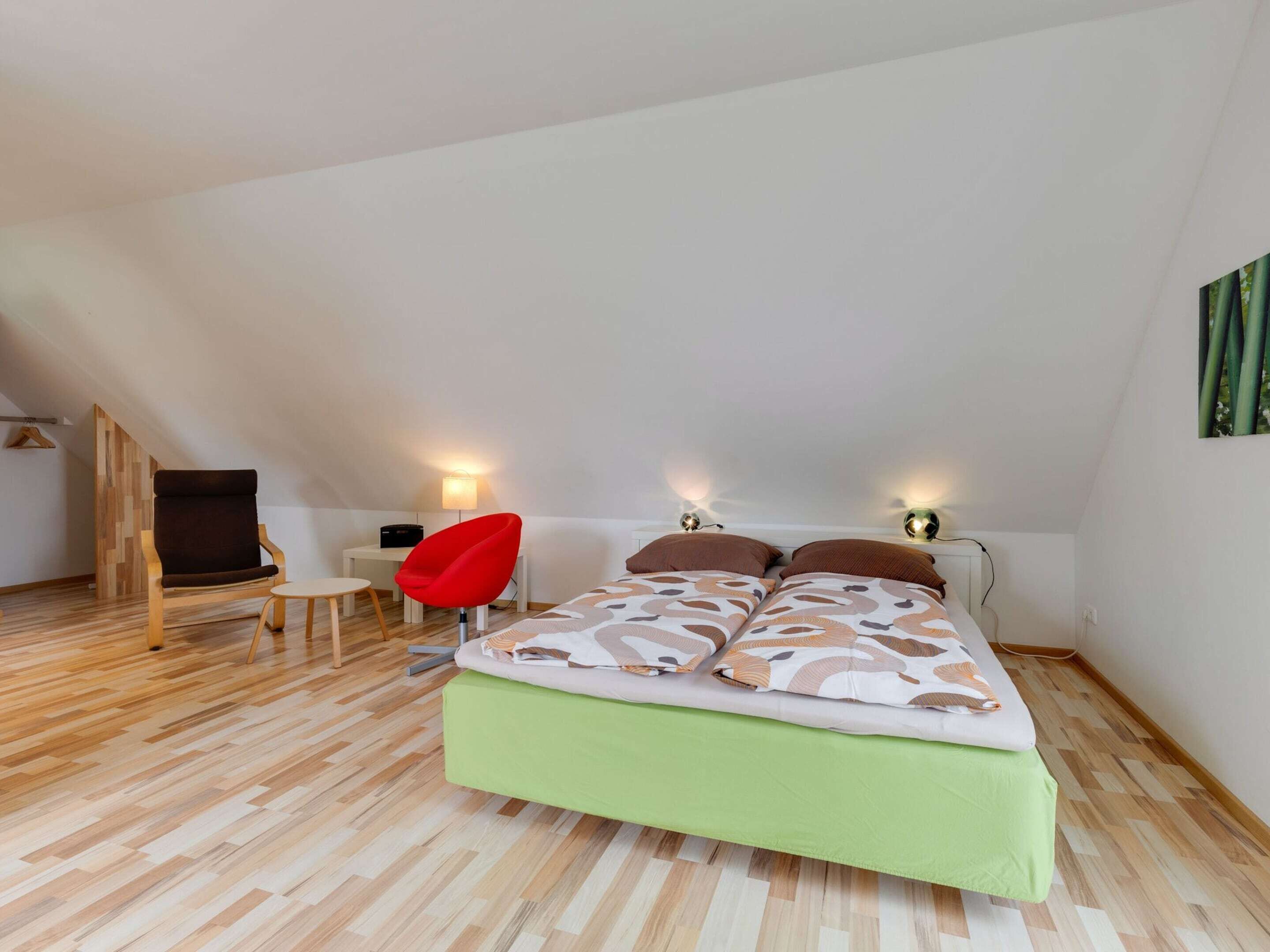 A bedroom with a loft-style ceiling, a bed with patterned bedding, a red chair, a small desk, and two wall-mounted lamps.