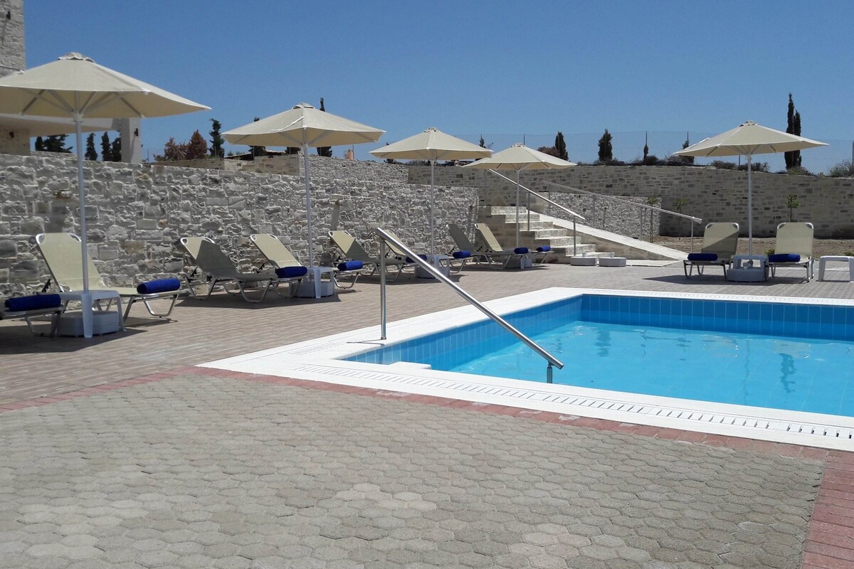 Un espace aménagé au bord de la piscine, avec des chaises longues et des parasols, un mur en pierre et un ciel d’un bleu limpide.