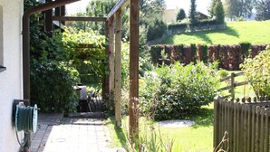 Un patio couvert équipé d’un enrouleur de tuyau d’arrosage, d’une pergola en bois et offrant une vue sur un jardin et une pelouse bien entretenus.