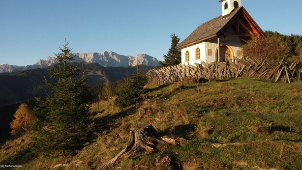 Une petite église dotée d’un clocher, située sur le versant d’une colline, entourée de montagnes et d’une clôture en bois.