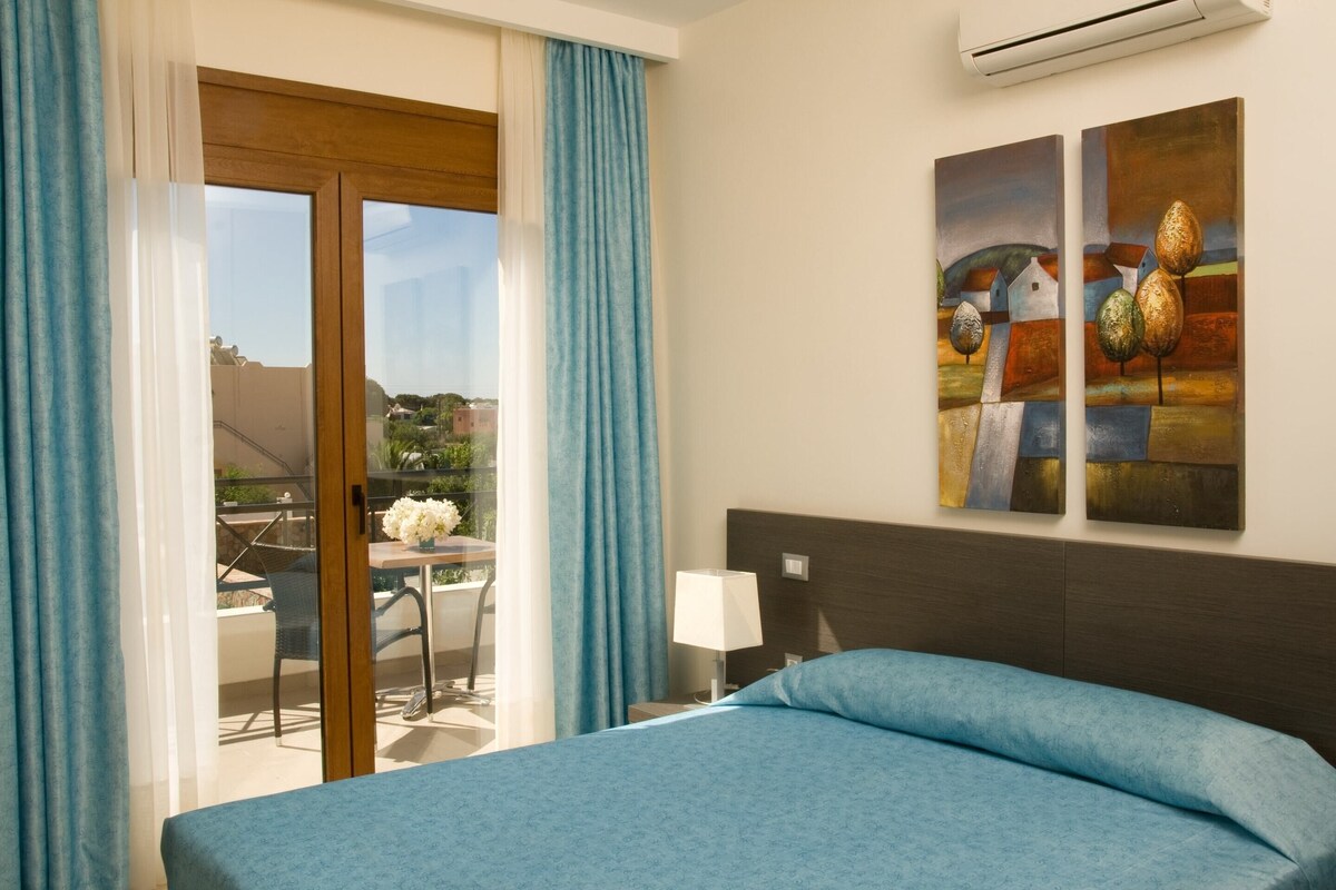 A hotel room with a bed, a view through a glass door to an outdoor area, and two paintings on the wall.