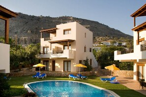A modern two-story building with a swimming pool, sun loungers, and a view of a mountainous landscape.