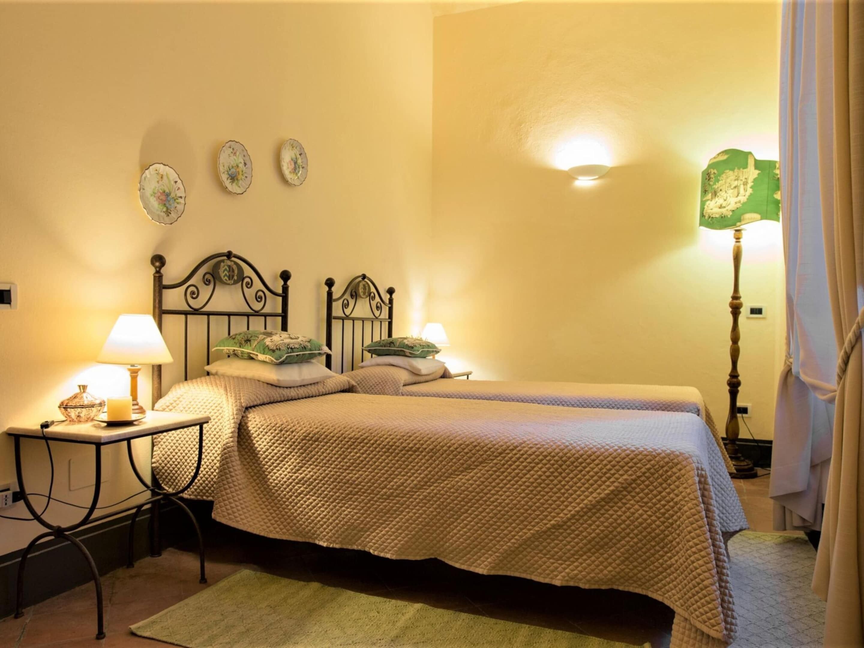 A bedroom with two single beds, a bedside table with a lamp, a green rug, and a wall-mounted lamp.