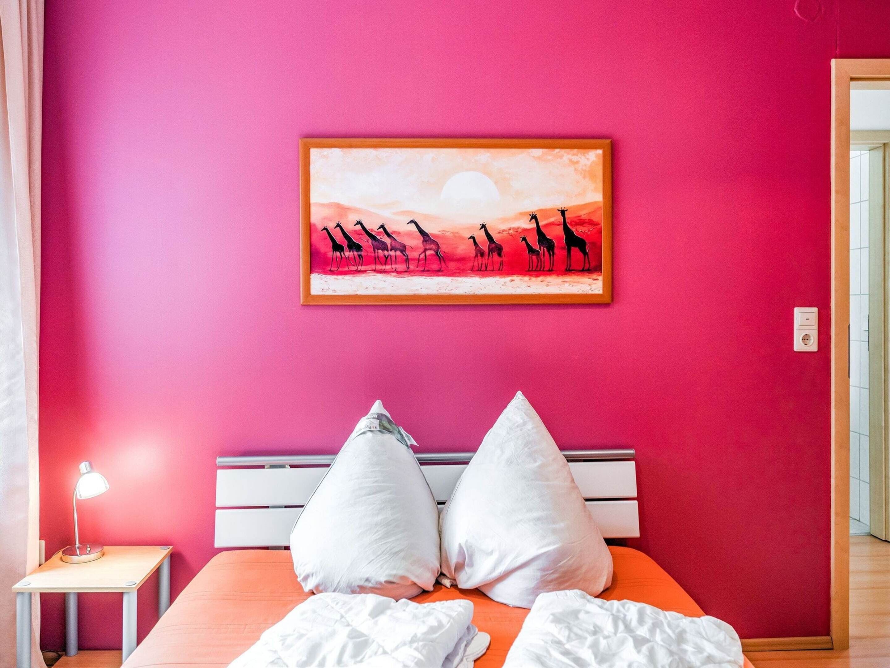 A bedroom with a pink wall, a framed painting of giraffes, a bed with white pillows, and a bedside table with a lamp.
