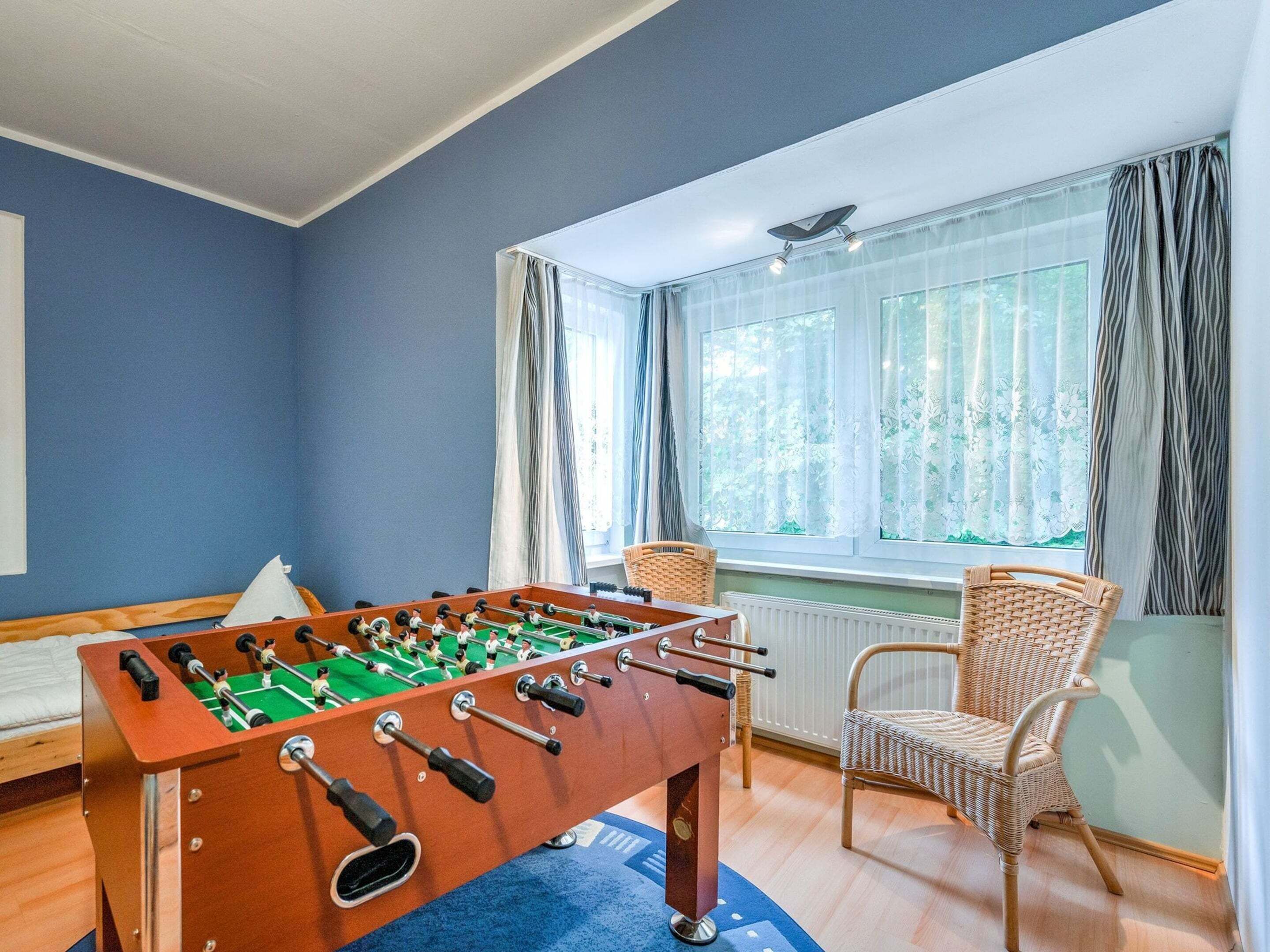 Una taula de futbolí a una habitació amb parets blaves, una cadira de vimet i una finestra amb cortines.