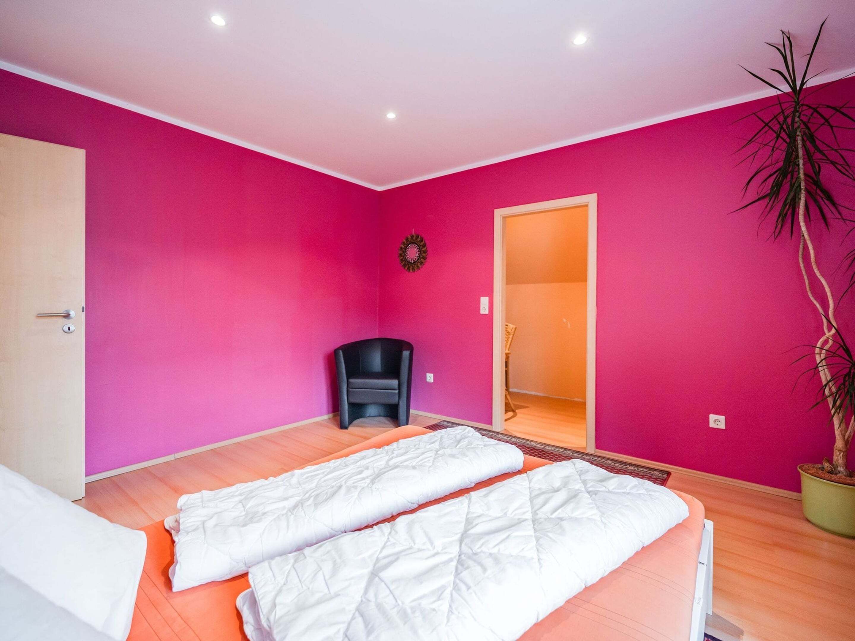 Ein Zimmer mit rosafarbenen Wänden, zwei Betten mit weißer Bettwäsche, ein schwarzer Sessel, ein Blumentopf und eine Tür zu einem anderen Raum.