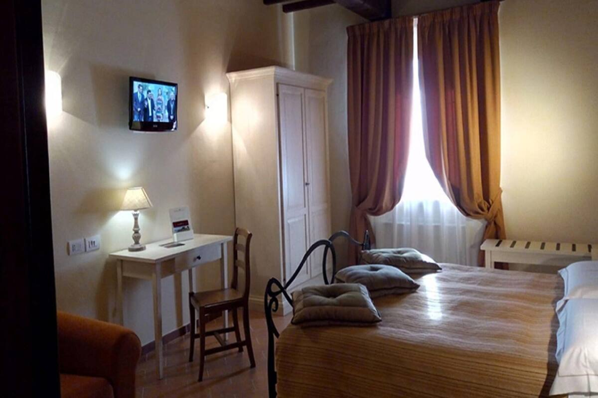 A bedroom with a wooden bed, a desk with a lamp, a chair, and a television on the wall.
