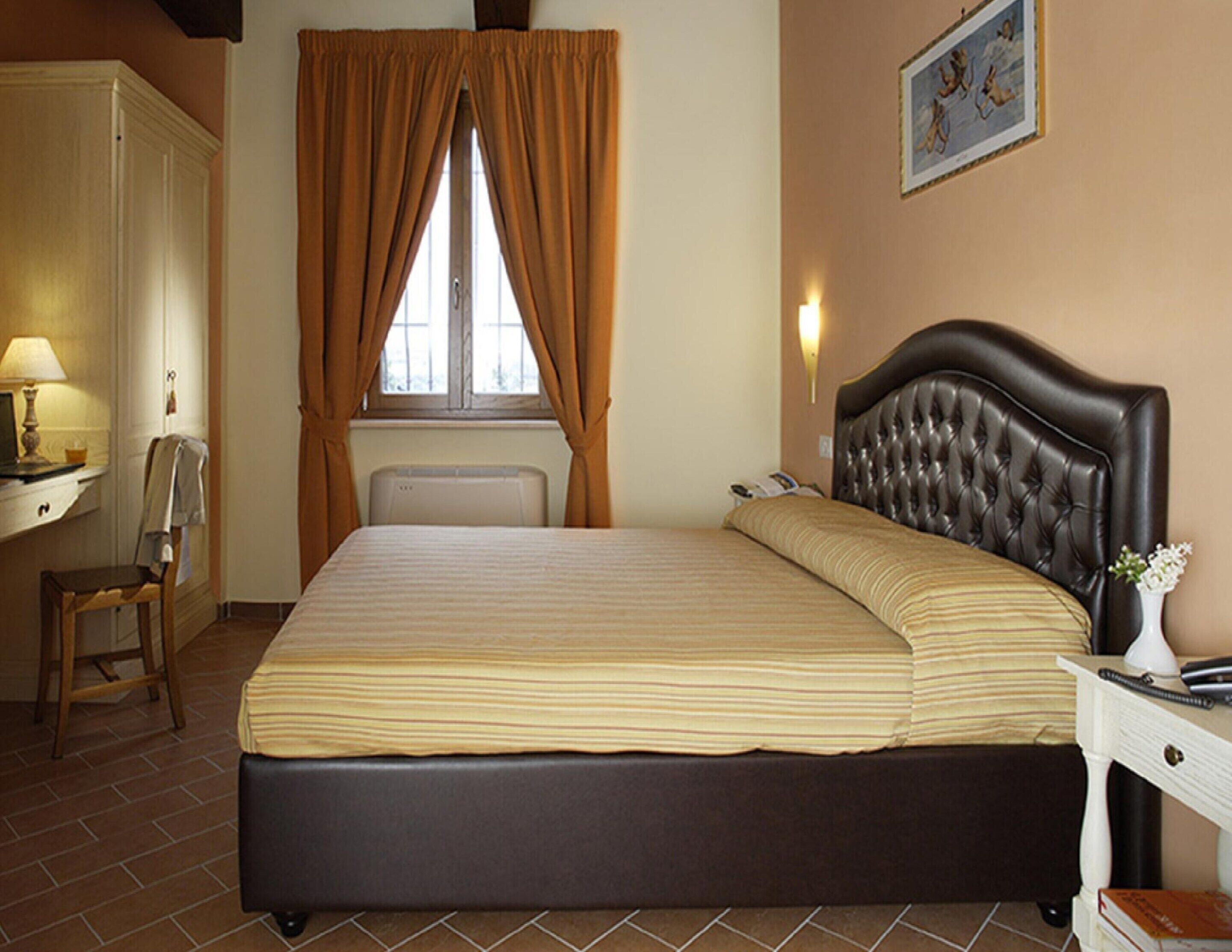 Habitación de hotel con una cama grande, un escritorio con lámpara, una silla, un radiador y un cuadro en la pared.