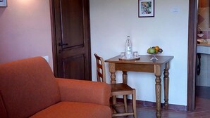 Ein Wohnzimmer mit einer Couch, einem Holzesstzimmer mit Stühlen, einem kleinen Tisch mit einer Wasserflasche und Obst, und einem gerahmten Bild an der Wand.