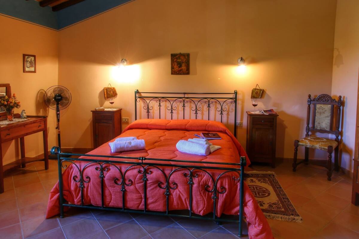 Ein Schlafzimmer mit einem Metallbett, zwei Nachttischen mit Lampen, einem Sessel und einem Ventilator.