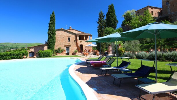 Une piscine avec des chaises longues et des parasols, un bâtiment en briques et un ciel d’un bleu limpide.
