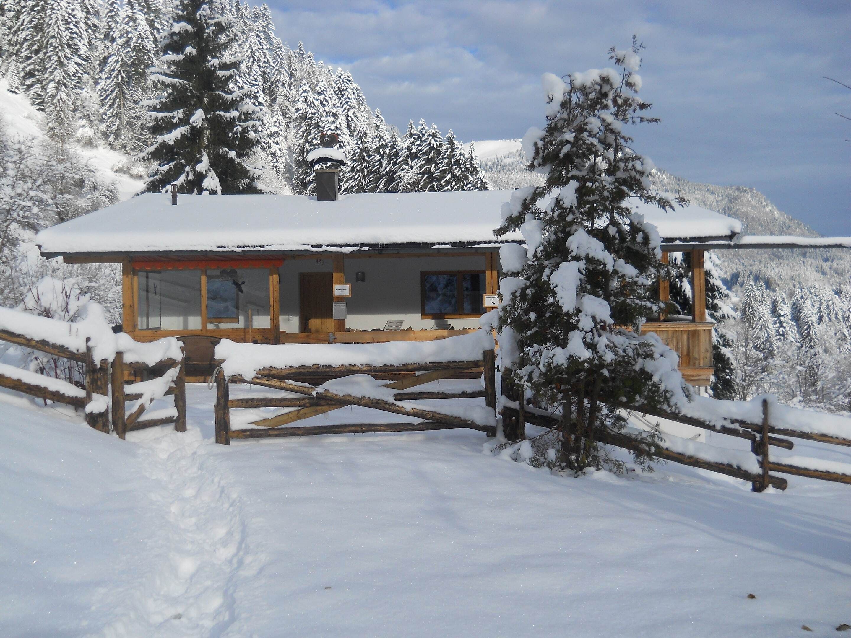 A wooden cabin with a snow-covered roof, surrounded by snow-laden trees and a mountain backdrop.