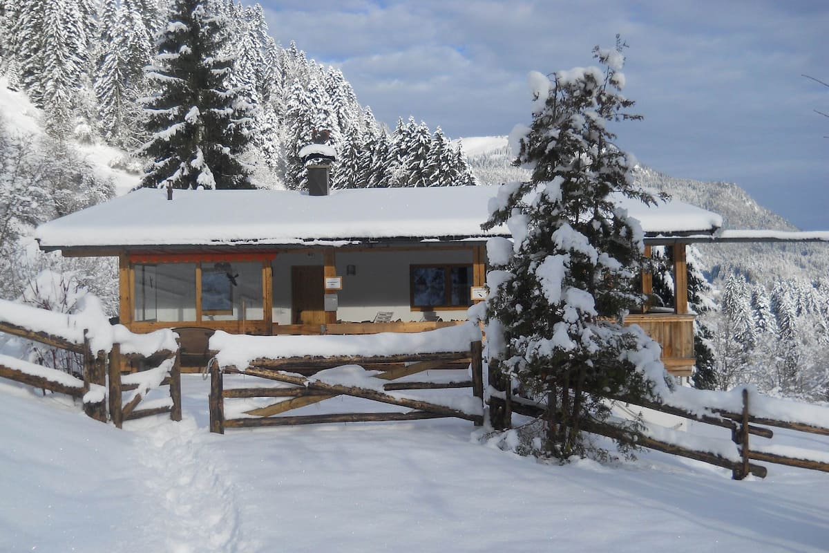 Una cabaña de madera con techo cubierto de nieve, rodeada de árboles cargados de nieve y una valla de madera.