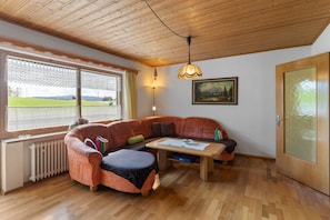 A cozy room with a wooden ceiling, a large window with lace curtains, a sectional sofa, a small table, and a door with frosted glass. - Cozy Apartment in Sonnen Bavaria Near Forest (Sonnen)