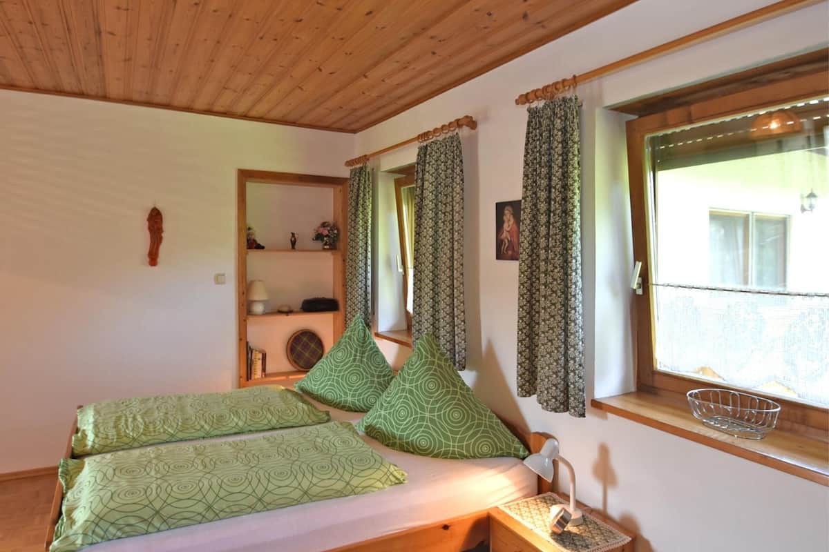 A room with a wooden ceiling, a bed with green patterned bedding, a nightstand with a lamp, and a window with patterned curtains.
