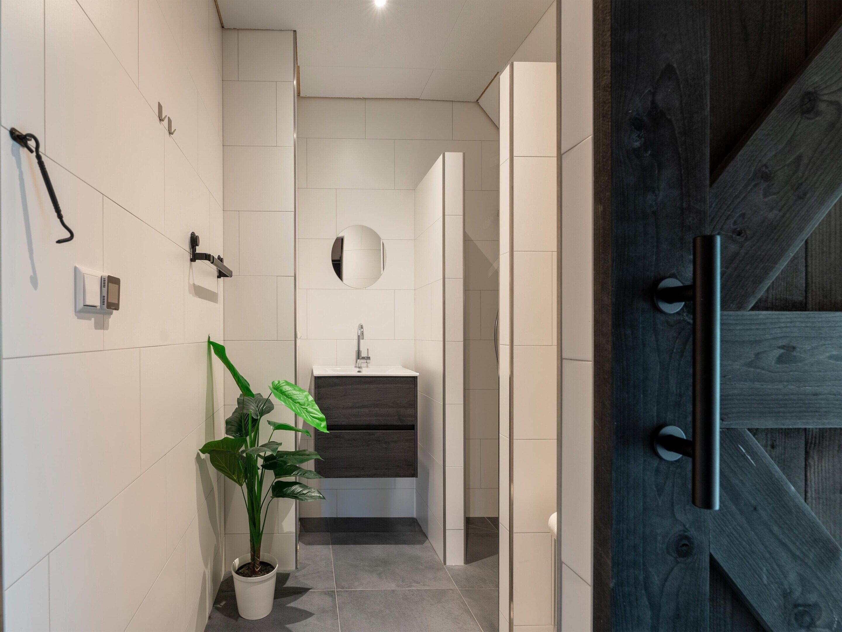 A modern bathroom with a white tiled wall, a round mirror, a sink, and a potted plant.