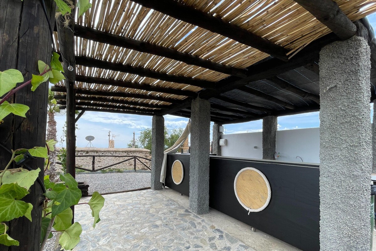 A covered outdoor area with stone flooring, wooden beams, and a view of a beach and wind turbines.