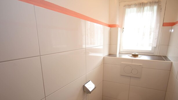 Un baño pequeño y limpio con azulejos blancos, un inodoro y una ventana con cortina.