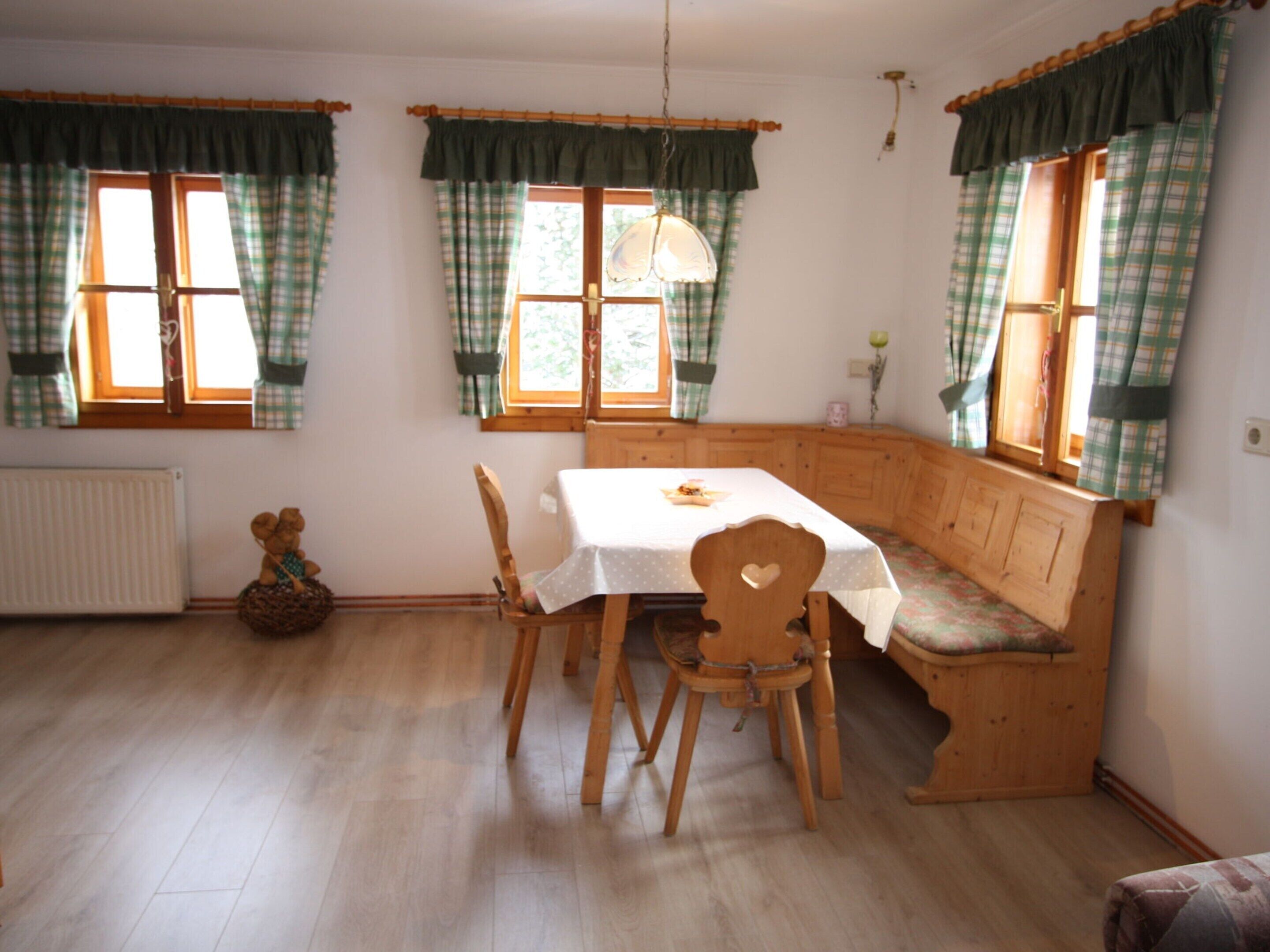 Een eetgedeelte met een houten bank, een tafel en stoelen. Er zijn ramen met gordijnen, een radiator en een teddybeer op de vloer.