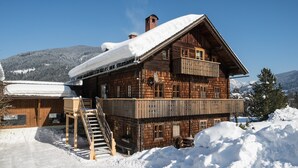 Eine Holzchalet mit Balkon, Treppe und verschneiter Landschaft im Hintergrund.