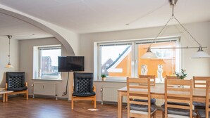 Un salón espacioso con piso de madera, un televisor montado en la pared y una zona de comedor con sillas y mesa de madera.
