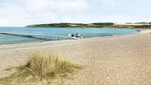 Ein Strand mit einem Steg, der sich in ruhiges Wasser erstreckt, einer Grünfläche und einer entfernten Hügelkette.
