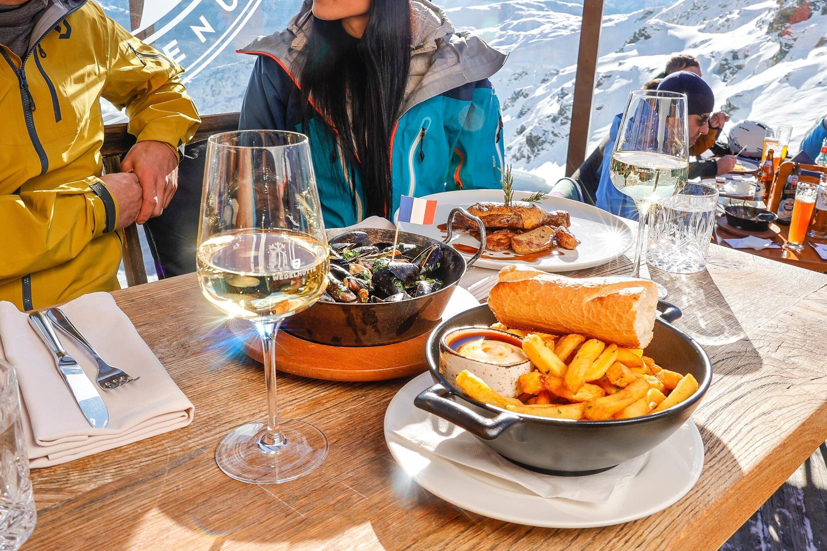 A dining table with a variety of dishes including mussels, fries, and a baguette, set against a snowy mountain backdrop.