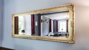 Una habitación con un espejo grande y decorativo en la pared, una cama, una ventana con cortinas y una lámpara sobre una mesita de noche.