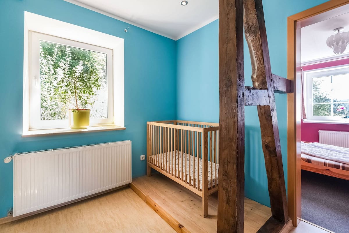 Una habitación con una cuna de madera, un ventanal con vistas a la vegetación y un radiador.