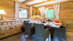 Interno di una baita in legno con zona pranzo, sedie in vimini, un armadietto in legno e un lampadario.