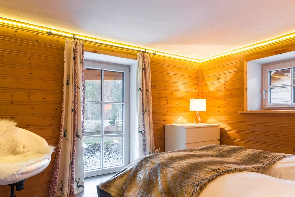 Una habitación acogedora con paredes de madera, una cama con una manta de piel sintética, una mesita de noche con una lámpara y una ventana con cortinas.