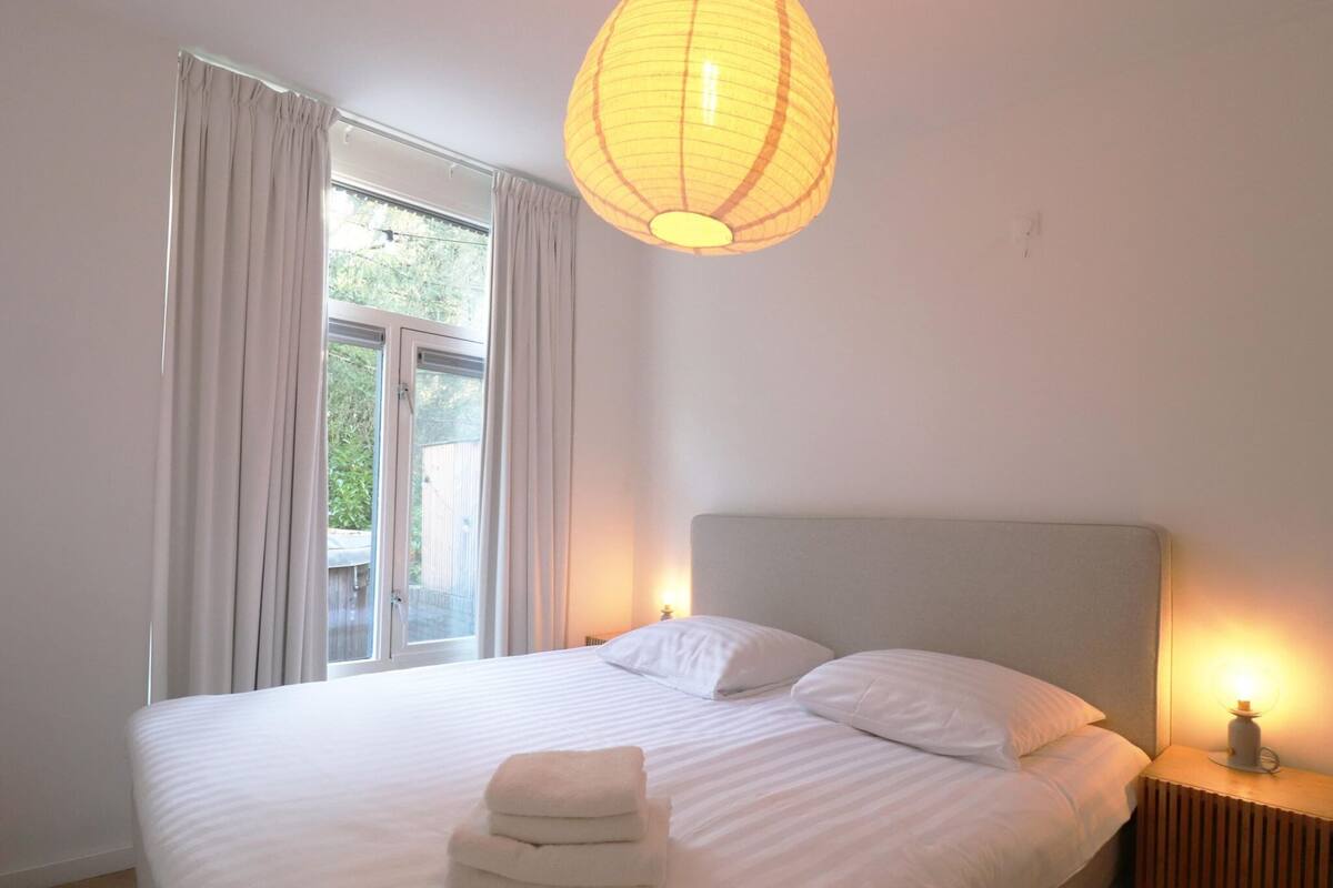 A neatly made bed with white linens and a folded towel, a bedside table with a lamp, and a window with curtains.