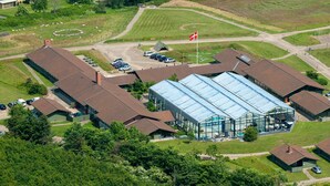 Eine Luftaufnahme eines Gebäudekomplexes mit mehreren Bauten, darunter eine große Glasgalerie, Parkplätzen und umliegender Bepflanzung.