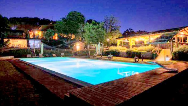 A swimming pool with a wooden deck, surrounded by well-lit buildings and trees. - Comfortable Cottages in Loro Ciuffenna (Terranuova Bracciolini)