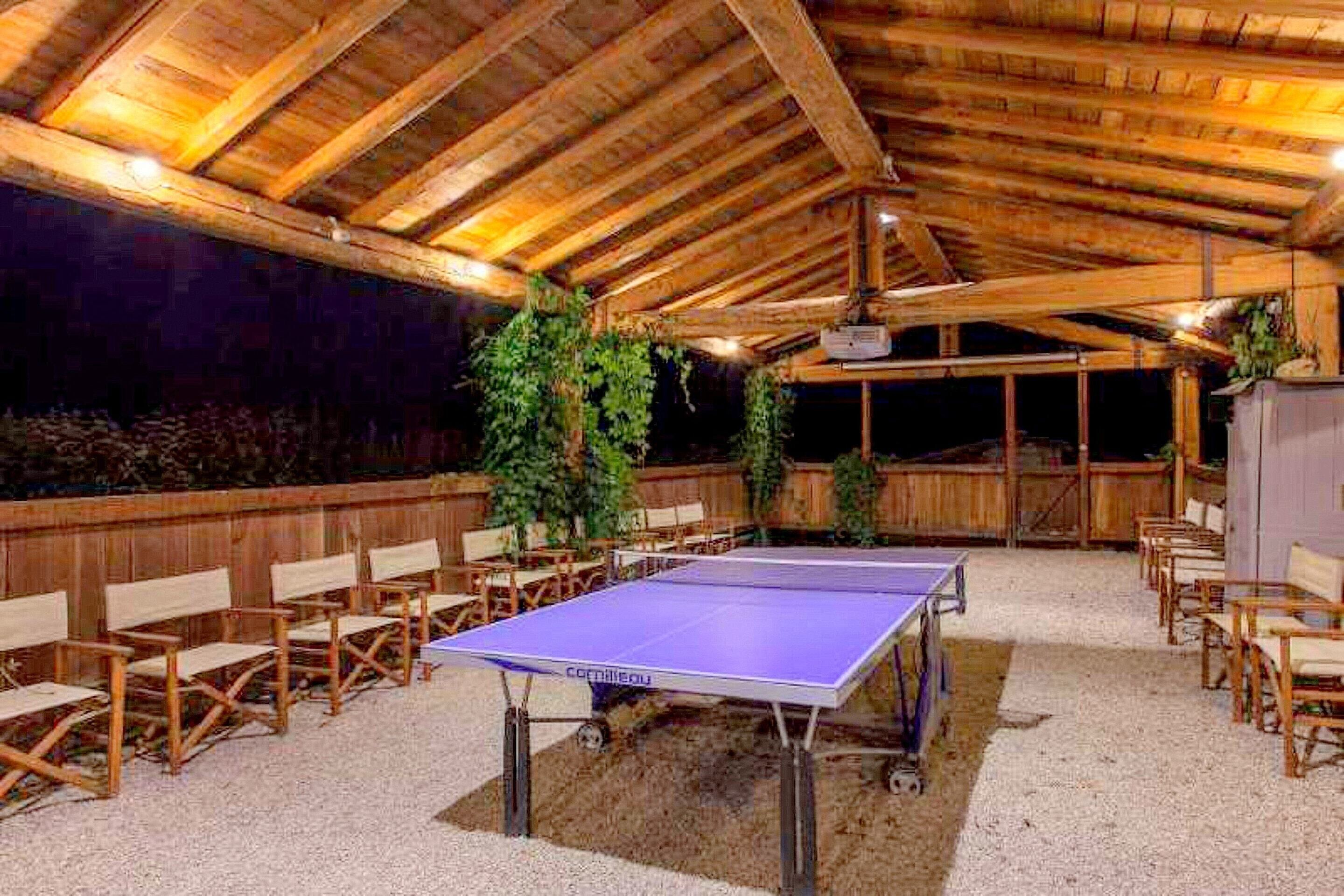 An outdoor ping pong table with surrounding chairs, a wooden ceiling, and a gravel floor.