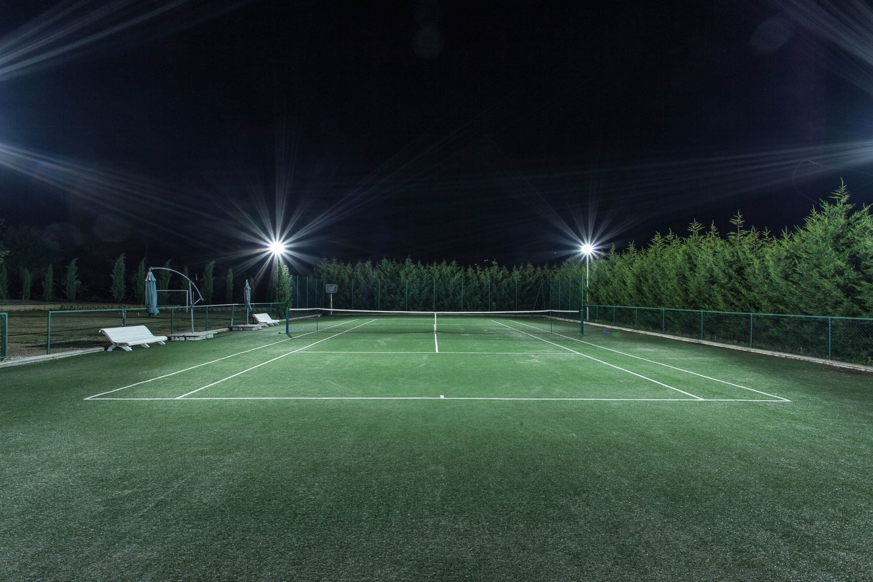 An outdoor tennis court at night with artificial lighting, surrounded by trees and a fence.