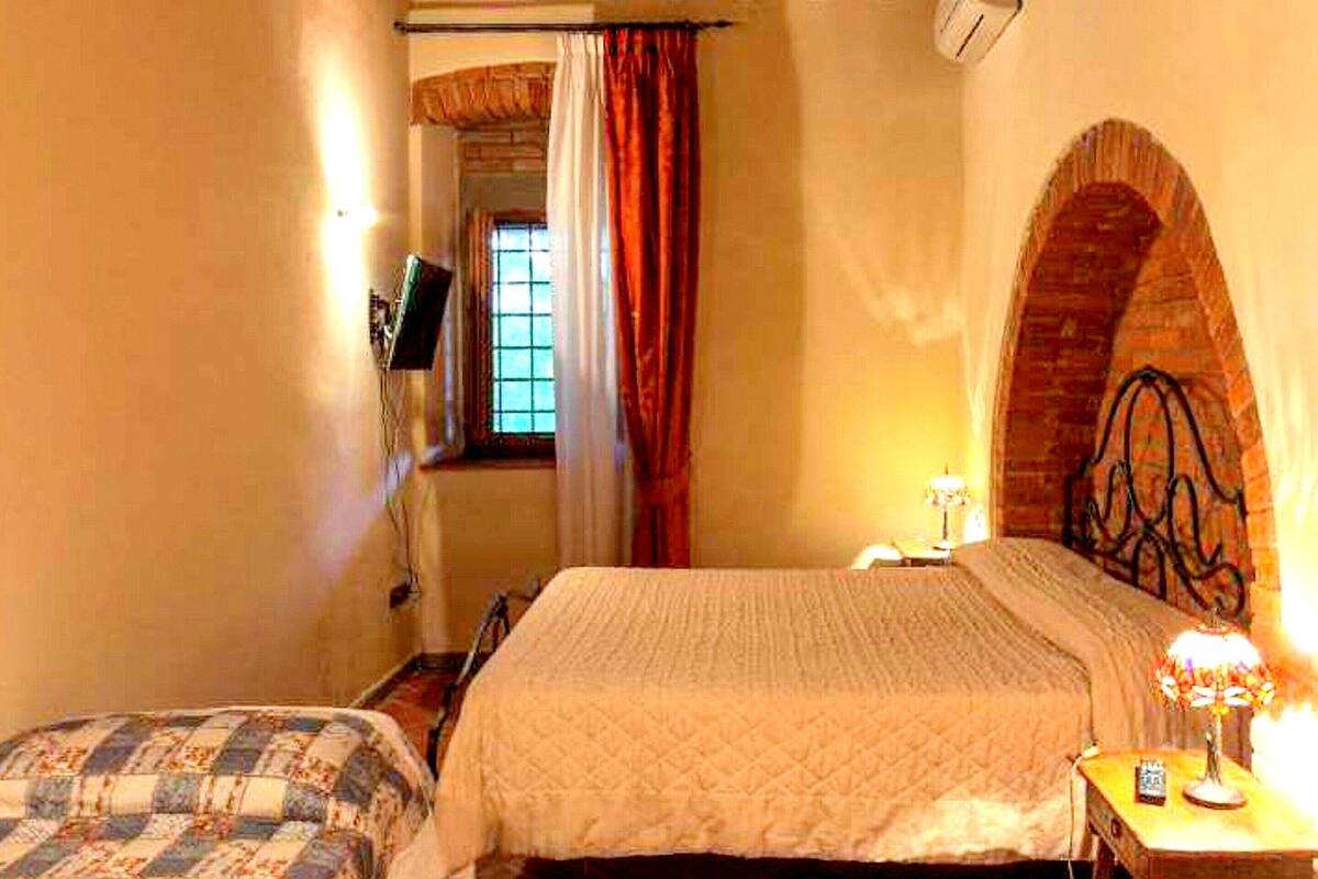 A room with a large bed, a smaller bed, a desk with a lamp, and a window with curtains.
