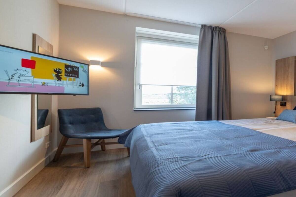 Egy modern szállodaszoba, amelyben egy nagy ágy, egy íróasztal és egy szék található. Az ablakból egy hídra nyílik kilátás, a falon pedig egy festmény látható.