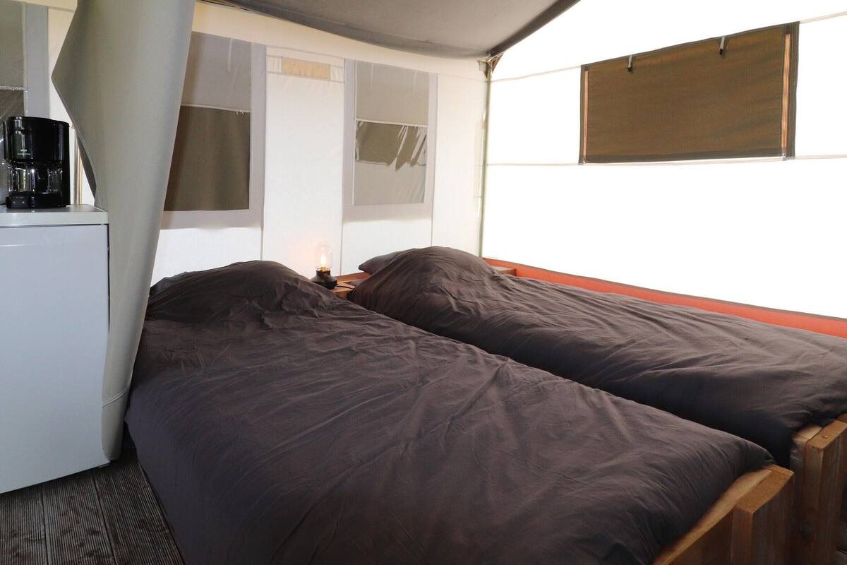 A small, enclosed room with two single beds, a desk with a coffee maker, and a window with a blind.