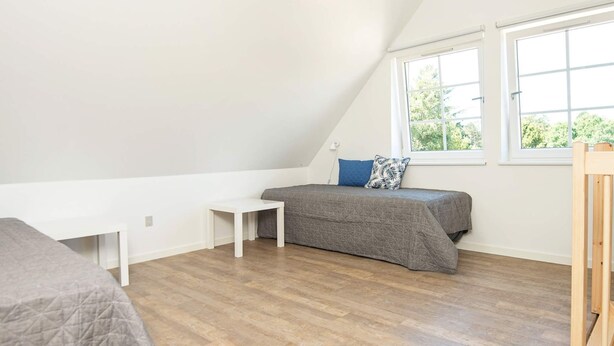 Una habitación pequeña y bien iluminada con techo inclinado, una cama con colcha gris, una mesita blanca y una ventana con vistas a la vegetación.