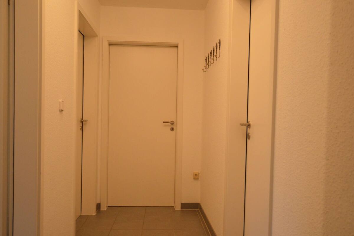 A narrow hallway with a closed door at the end, tiled floor, and a wall-mounted light fixture.