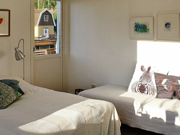 A bedroom with two beds, a window with a view of a house, and a lamp on the left side.
