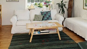 A living room with a white sofa, a wooden coffee table with books, a green patterned cushion, and a view of a marina through a large window. - 4 Person Holiday Home in Ellos (Ellos)